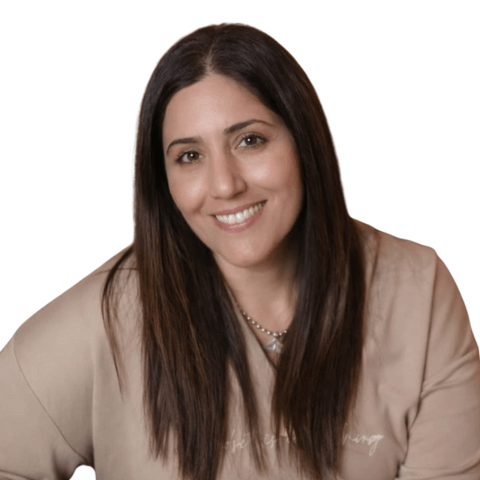 אישה מחייכת עם שיער חום כהה, חולצה בצבע בז' ותליון כסף.