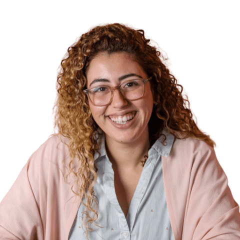 אישה חייכנית עם שיער מתולתל ומשקפיים, לובשת חולצה מכופתרת וקרדיגן ורוד.